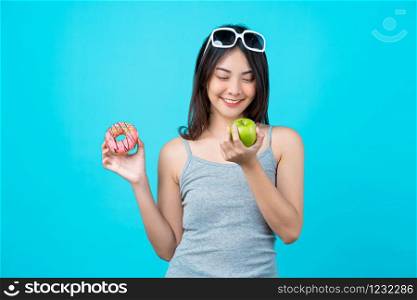 Attractive Asian young woman holding and choosing between disk of donuts or green apple fruit on isolated blue color background, weight loss and avoid junk food for dieting and Healthy