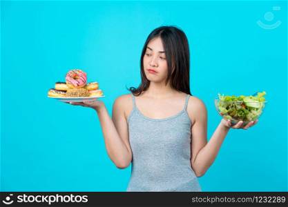 Attractive Asian young woman holding and choosing between disk of donuts or vegetable salad in glasses bowl on isolated blue color background, weight loss and avoid junk food for dieting and Healthy