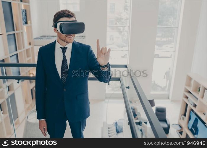 Attractive bearded businessman in formal black suit wears wireless VR headset goggles, surfing in virtual reality while standing in company lobby. Digital future and innovation technology concept. Attractive bearded businessman in formal black suit wears wireless VR headset goggles