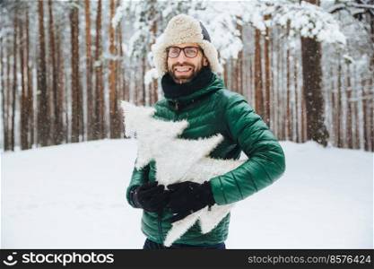 Attractive bearded male with appealing appearance, wears green anorak and hat, holds artificial fir tree, spends weekends on frosty weather in winter forest. Winter, weather and lifestyle concept