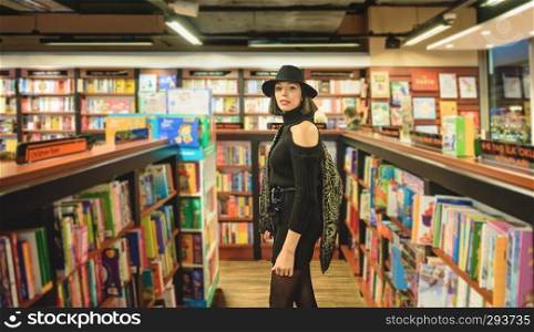 Attractive beautiful woman with black hat and dress stands in bookstore while looking towards the camera