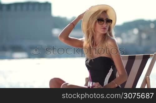Attractive blond female in hat sitting on sun lounger on the beach and posing while drinking cocktail