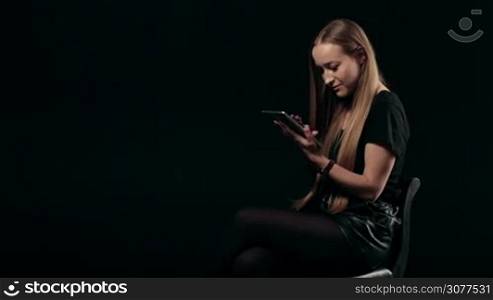 Attractive blonde female with amazing long hair sitting on chair and working with digital tablet computer isolated on black background. Young woman surfing the net with touchpad and looking at screen.