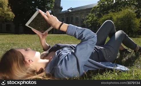 Attractive blonde girl lying on her back with touchpad on green grass while studying on campus. Closeup. Dolly Shot. College female student surfing the net with digital tablet pc with blurry group of university friends communicating on the background