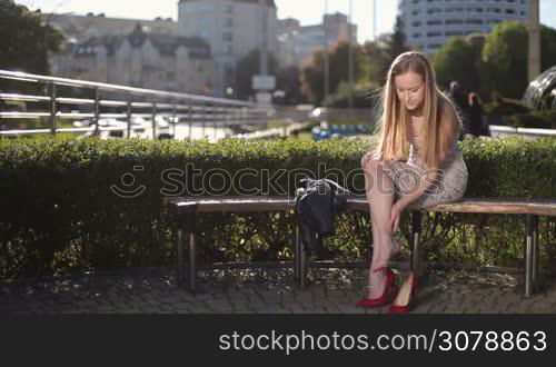 Attractive blonde woman in red high heels massaging tired legs while sitting on the bench in the park. Sensual young female in high heel shoes suffering from leg pain, massaging aching feet and touching her ankle while relaxing outdoors.