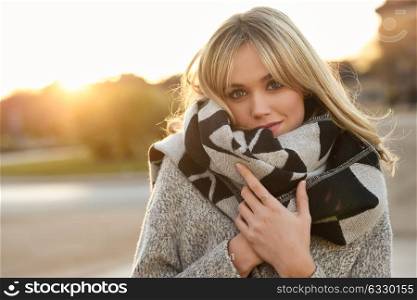 Attractive blonde woman in urban background with sun backlight. Young girl wearing winter coat and scarf standing in the street. Pretty female with straight hair hairstyle and blue eyes.
