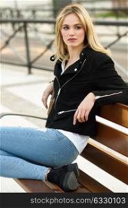 Attractive blonde woman in urban background. Young girl wearing black zipper jacket and blue jeans trousers sitting on a bench in the street. Pretty female with straight hair hairstyle and blue eyes.