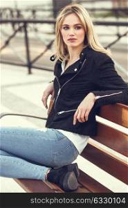 Attractive blonde woman in urban background. Young girl wearing black zipper jacket and blue jeans trousers sitting on a bench in the street. Pretty female with straight hair hairstyle and blue eyes.