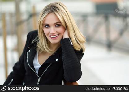 Attractive blonde woman smiling in urban background. Young girl wearing black zipper jacket sitting on a bench in the street. Pretty female with straight hair hairstyle and blue eyes.