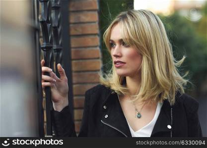 Attractive blonde woman standing in urban background with lost look. Young girl wearing black zipper jacket. Pretty female with straight hair hairstyle and blue eyes.