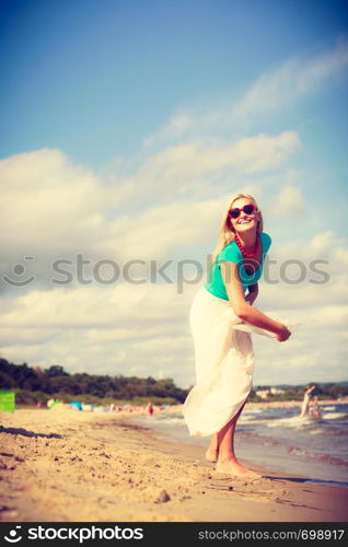 Attractive blonde woman wearing long romantic dress walking on beach and having fun during summer, throwing sun hat.. Woman on beach throwing sun hat