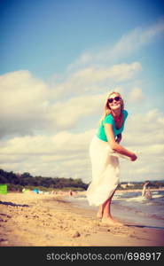 Attractive blonde woman wearing long romantic dress walking on beach and having fun during summer, throwing sun hat.. Woman on beach throwing sun hat