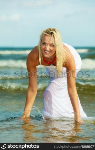 Attractive blonde woman wearing long white romantic dress playing with water in sea during summer.. Blonde woman wearing dress playing with water