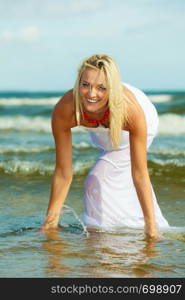 Attractive blonde woman wearing long white romantic dress playing with water in sea during summer.. Blonde woman wearing dress playing with water