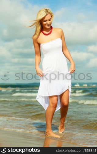 Attractive blonde woman wearing long white romantic dress walking in water sea during summer.. Blonde woman wearing dress walking in water