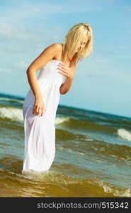 Attractive blonde woman wearing long white romantic dress walking in water sea during summer.. Blonde woman wearing dress walking in water