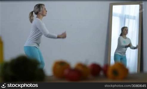 Attractive blonde woman with ponytail doing crisscross crunches near mirror at home. Sporty middle-aged female working out in domestic room with blurry vegetables on foreground. Healthy lifestyle.