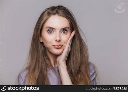 Attractive blue eyed brunette female looks with bewilderment at camera, has specific appearance, can`t beleive in eyes, isolated over grey studio background. People and facial expressions concept