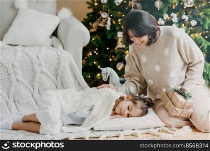 Attractive brunette smiling woman stands on knees with present boxes, little daughter sleeps near on pillow, has pleasant dreams, enjoy holiday. Winter time, New Year and Christmas. Rest concept