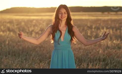 Attractive brunette woman in stylish blue dress with arms outstreched spinning around in glow of beautiful sunset. Young female enjoying nature, sunlight, freedom as she whirls around with eyes closed at sunset against golden wheat field background