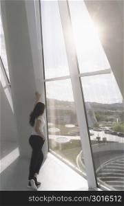 Attractive brunette woman, leaning against a wall, looking on large windows, under the direct sunlight.