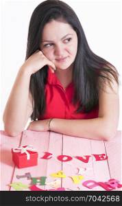 Attractive brunette woman thinking while sitting at a pink table, with a red gift and the word love spelled from red paper letters in front of her.