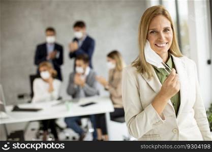 Attractive business woman taking off her protective facial mask and looking at the camera with her team members sitting in the background