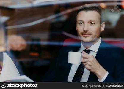 Attractive businessman in formal suit, drinks coffee during dinner break, sits in luxury cafe, looks thoughtfully at window, contemplates about business affairs has pleasant look. People and lifestyle