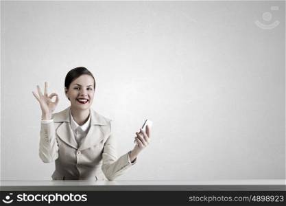 Attractive businesswoman having mobile conversation. Young pretty businesswoman sitting at table with mobile phone in hand showing ok gesture