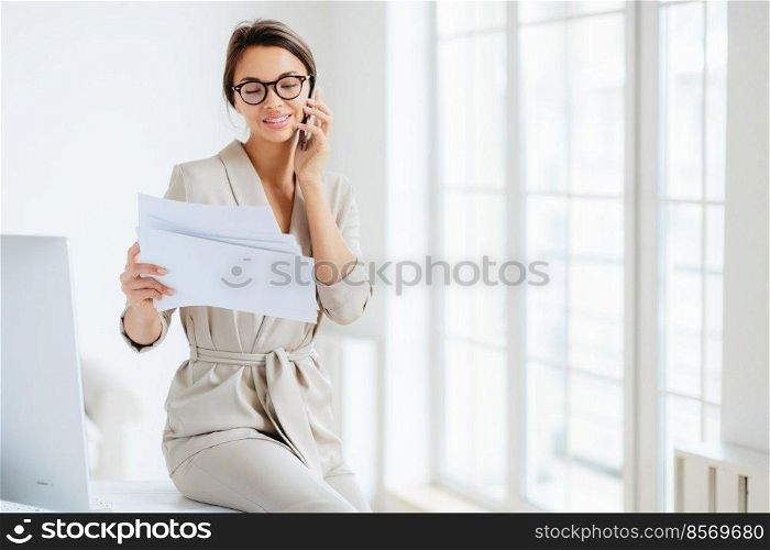 Attractive businesswoman reads papers or business documents, has telephone talk with business partner, examines paperwork before meeting, sits at desktop, smiles happily, works with documentation