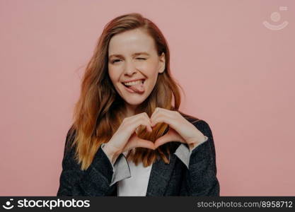 Attractive carefree young woman with wavy ginger hair in formal dark jacket showing heart shape sign, winking and sticking tongue out standing against light pink wall in studio, expressing fun. Happy excited young woman showing heart shape sign