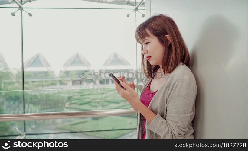 Attractive casual happy young Asian woman in international airport, using and checking her smartphone standing in terminal hall while waiting for her flight to board airplane at the departure gate.
