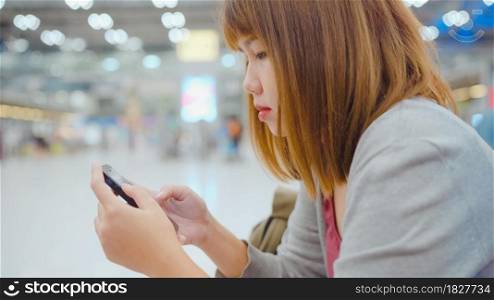 Attractive casual happy young Asian woman in international airport, using and checking her smartphone standing in terminal hall while waiting for her flight to board airplane at the departure gate.