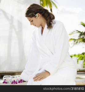Attractive Caucasian mid-adult woman at spa wearing white robe sitting looking down at candles and orchids.