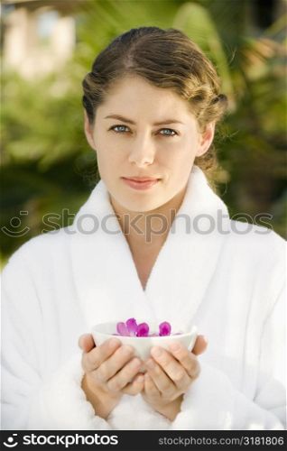 Attractive Caucasian mid-adult woman in white robe holding bowl of purple orchids floating in water.