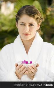Attractive Caucasian mid-adult woman in white robe holding bowl of purple orchids floating in water.