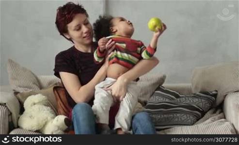 Attractive caucasian mother and her curly mixed race toddler boy sitting on the sofa and playing. Loving mother and adorable little son with apple in his hand spending leisure and having fun at home. Slow motion.