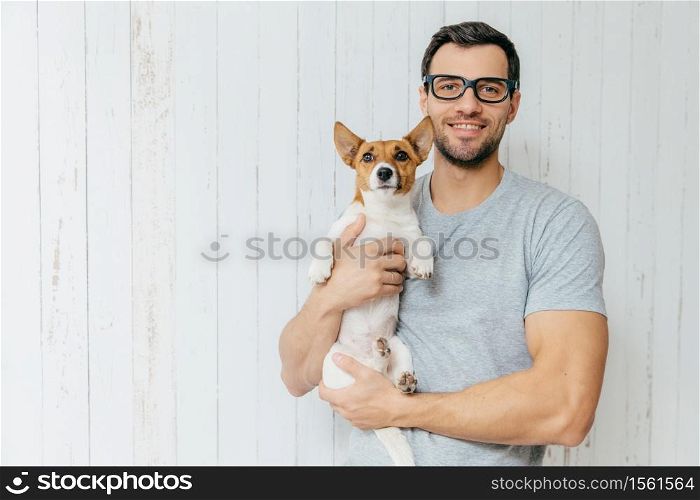 Attractive cheerful Caucasian male in casual t shirt, spectacles, holds favourite pet, stands against white wooden background with blank space. Happy unshaven man with his jack russell terrier