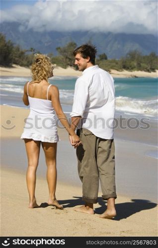 Attractive couple holding hands and smiling at eachother walking on Maui, Hawaii beach.