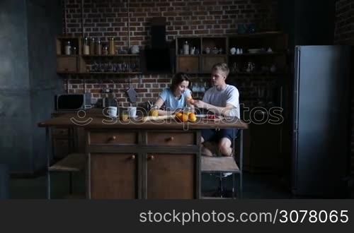 Attractive couple in love sitting at kitchen table and having breakfast together in the morning. Affectionate man feeding his cute girlfriend with tasty croissant. Young family enjoying meal together in modern kitchen.