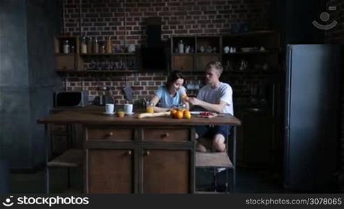 Attractive couple in love sitting at kitchen table and having breakfast together in the morning. Affectionate man feeding his cute girlfriend with tasty croissant. Young family enjoying meal together in modern kitchen.