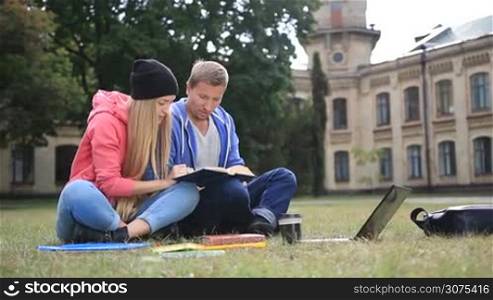 Attractive couple of students sitting on the grass and reading a book outside the college in the park. Young smiling students studiyng together outdoors.
