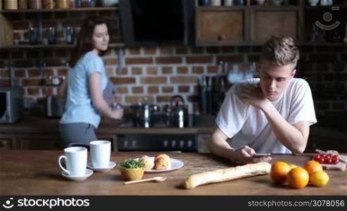Attractive couple relaxing in the kitchen in the morning. Cute young woman bringing orange juice, hugging her boyfriend and kissing him with tenderness while handsome hipster browsing social networks with mobile phone.