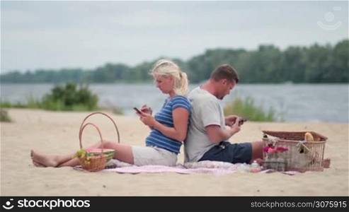 Attractive couple sitting back to back on the beach and texting messages on cell phones during summer holidays