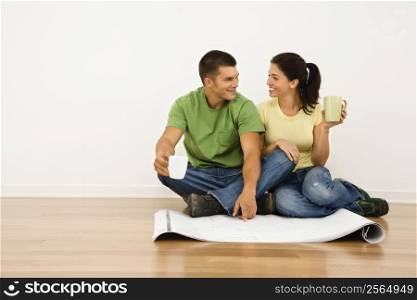 Attractive couple sitting on home floor with coffee cups looking at house plans and smiling at eachother.