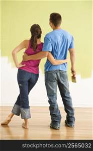 Attractive couple standing in front of partially painted wall with arms around eachother.