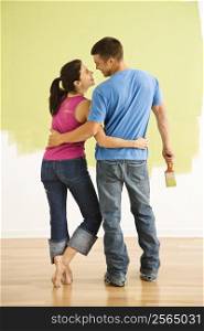 Attractive couple standing in front of partially painted wall with arms around eachother smiling.