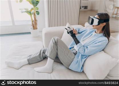 Attractive disab≤d girl is relaxing in vr glasses on couch. Handicapped caucasian person≥ts rehabilitation for sensor cyber arm. Amputee is setting functions of myoe≤ctric robotic limb.. Disab≤d girl relaxing in vr glasses on couch. Handicapped person setting functions of cyber arm.