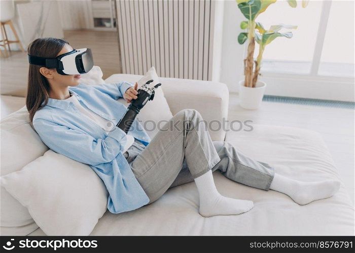 Attractive disabled girl is relaxing in vr glasses on couch. Handicapped caucasian person gets rehabilitation for sensor cyber arm. Amputee is setting functions of myoelectric robotic limb.. Disabled girl relaxing in vr glasses on couch. Handicapped person setting functions of cyber arm.
