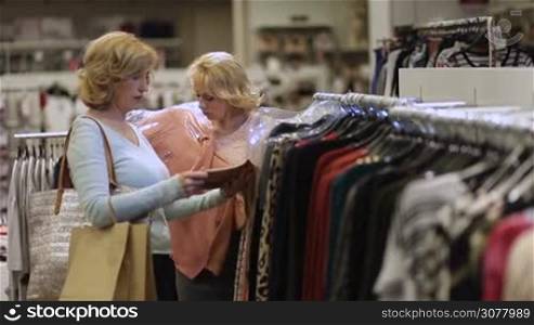 Attractive eldery females with shoulder bags looking at clothes hanging on hangers inside clothing store. Beautiful women selecting apparel while shopping in boutique fashion shop.